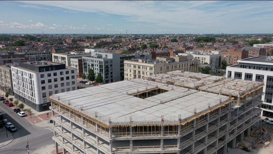 Luftaufnahme eines Rohbaus im dicht bebauten Stadtgebiet: Geschossdecken in Schalung, Bewehrung an den Rändern.